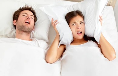 A woman covering her ears with a pillow while a man lies next to her and snores